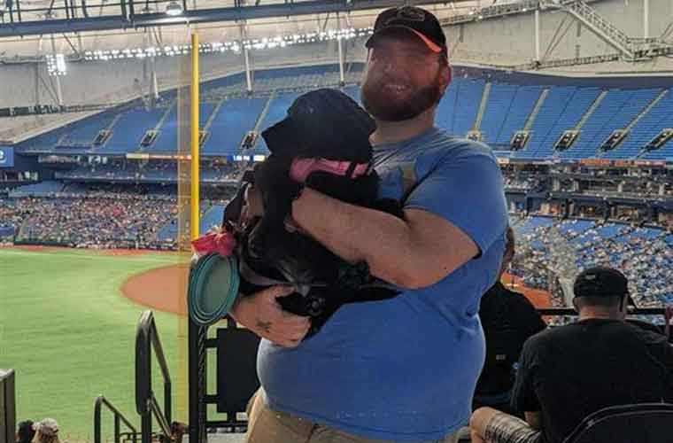 A tall man with a mustache and beard wearing a short-sleeved shirt and shorts holds a black dog in a sports stadium.