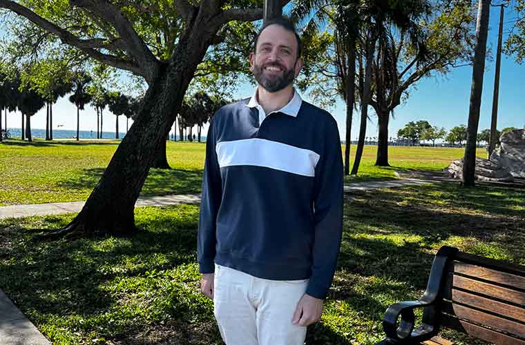 A man with a mustache and beard wears a long-sleeved dark blue shirt with a white stripe and white pants and stands in a waterfront park near a bench.