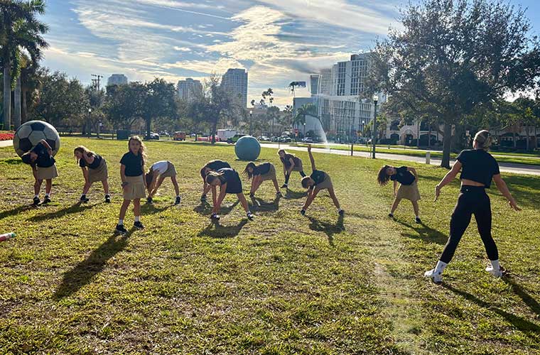 Several girls work with a woman in a black T-shirt and black pants on exercises and stretching 
