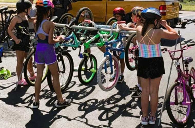 A group of little girls in shorts, shirts and bike helmets work with bikes that are attached to a rack in a park.