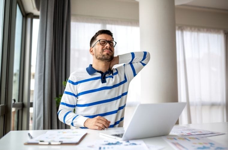 Man wearing glasses and a striped shirt stretches his neck while sitting at a desk working on a laptop, suggesting discomfort from poor posture.