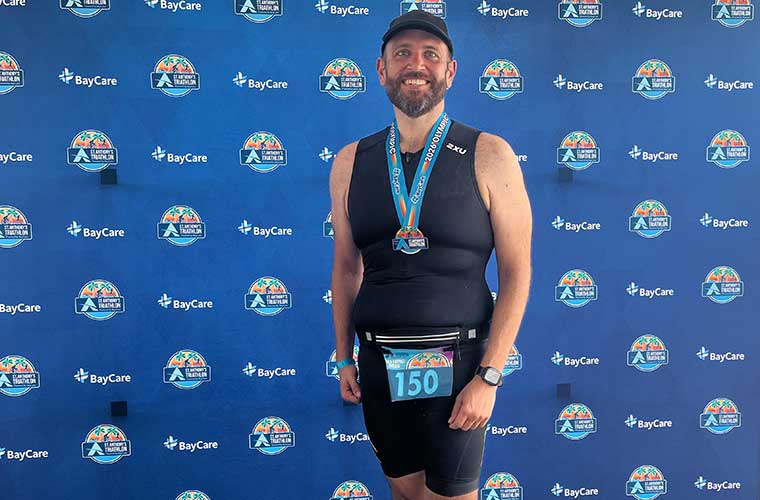 Man wearing athletic gear stands smiling against a backdrop displaying BayCare and St. Anthony's Triathlon logos. They are wearing a race bib numbered 150 and a medal around their neck.