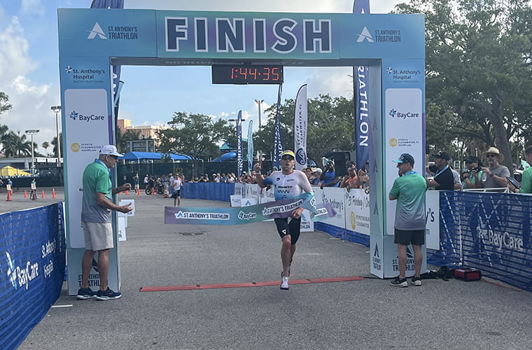 A person is crossing the finish line at a triathlon race, with a time displayed above. The event appears to be sponsored by entities like AdventHealth and BayCare, as seen on banners. Spectators and staff can be seen in the background.