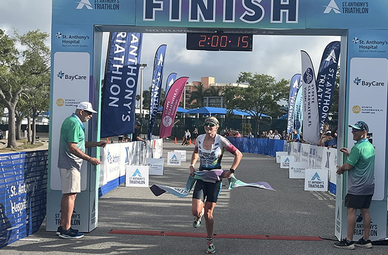 An athlete crosses the finish line at the St. Anthony's Triathlon. The digital timer displays 2:00:15. Staff members and spectators stand nearby under a sunny sky. Several banners and sponsor logos, including St. Anthony's Hospital and BayCare, are visible.