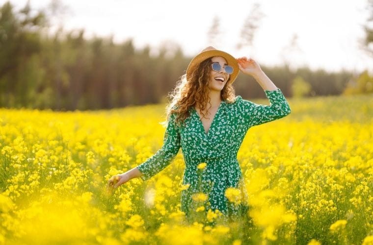 A person in a green patterned dress and hat stands in a field of yellow flowers, holding the brim of the hat.