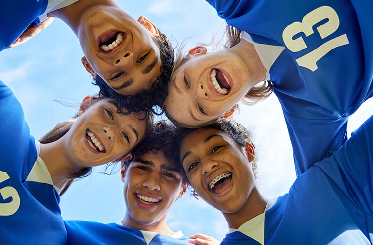 A group of people wearing blue soccer jerseys stand in a huddle, smiling and looking down at the camera. The sky is visible above them.