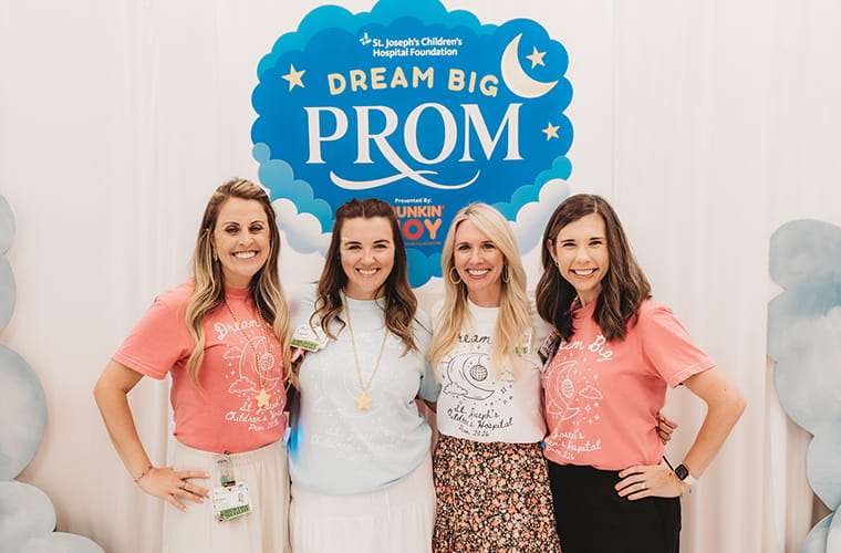 Four women stand smiling in front of a "St. Joseph's Children's Hospital Foundation Dream Big Prom" backdrop. They are wearing colorful shirts featuring prom-related designs.
