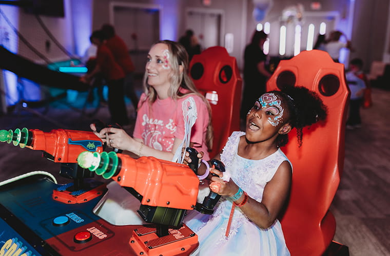 A woman and young girl sit in red gaming chairs, engaged in an arcade game with large orange and green controllers. The woman wears a pink shirt, and the girl is smiling with face paint and a light-colored dress. The background shows a softly lit room with other people and gaming equipment.