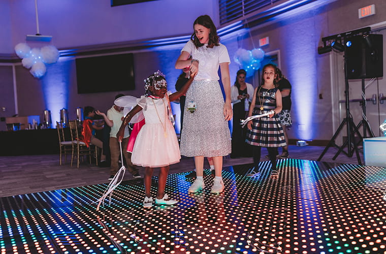 A woman and a young girl dance on an illuminated floor at a special event. The girl wears a light-colored dress with a fairy costume, including wings and a headpiece. Another child stands nearby, wearing a plaid dress. The atmosphere is festive, with blue lighting and decorative balloons in the background.