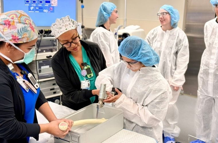 A BayCare career camp participant wearing personal protective equipment receives hands-on instruction from surgical staff in an operating room setting. The participant examines medical equipment while other camp attendees observe nearby.
