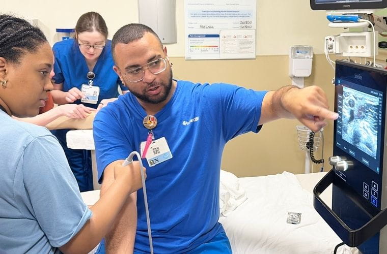 A BayCare career camp participant practices ultrasound imaging under the guidance of a clinical team member in a hospital training room. The participant holds an ultrasound probe while viewing images displayed on a monitor as additional staff members observe in the background.