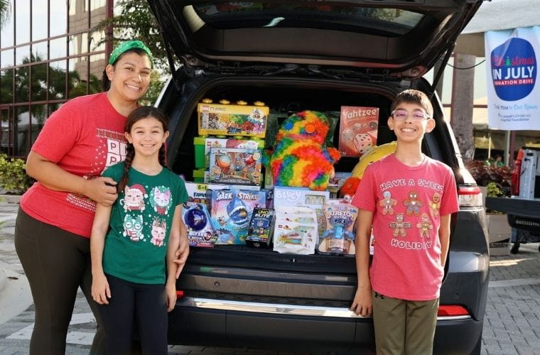 A person and two children stand beside the open trunk of an SUV filled with donated toys, games and stuffed animals. Visible items include board games, a colorful plush toy and several boxed children's toys. A banner for a Christmas in July donation drive is visible in the background near an office building.