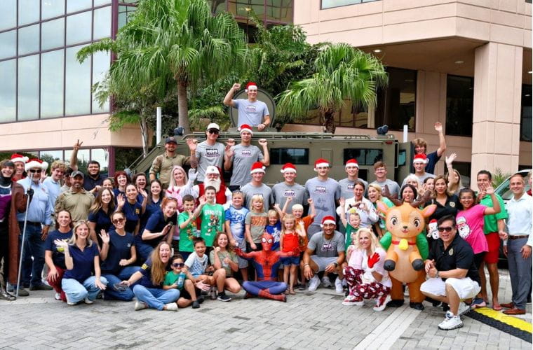 A large group of community members, volunteers, children and event supporters pose outdoors in front of a military-style vehicle during a Christmas in July celebration. Several participants wear Santa hats, and holiday-themed characters are visible in the group.