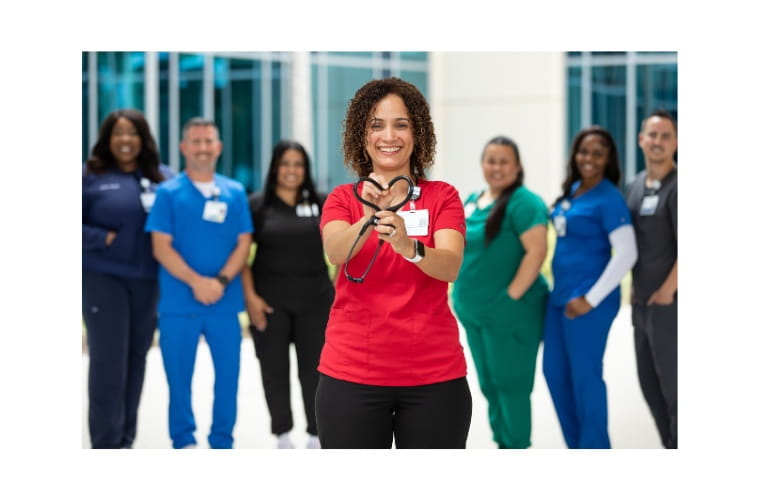 Surrounded by health care team members in scrubs of varying colors, a smiling BayCare team member holds a stethoscope in a heart shape in front of her.