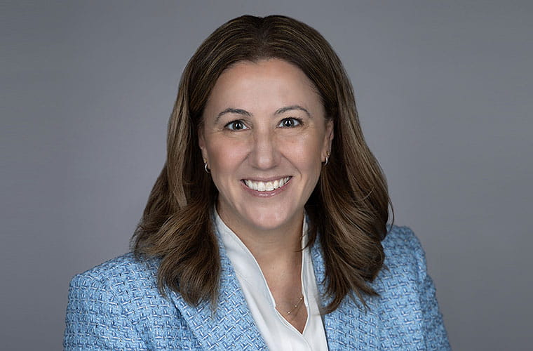 A woman with brown hair and wearing a white blouse and blue jacket smiles into the camera.