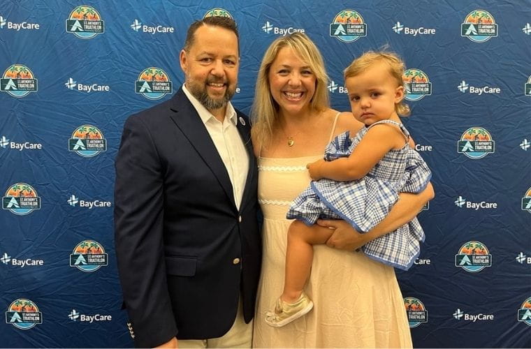 A man wearing a white shirt and a blue blazer stands with a woman in a tan dress who is holding a little girl in a blue checked dress in front of a blue background with logos on it.