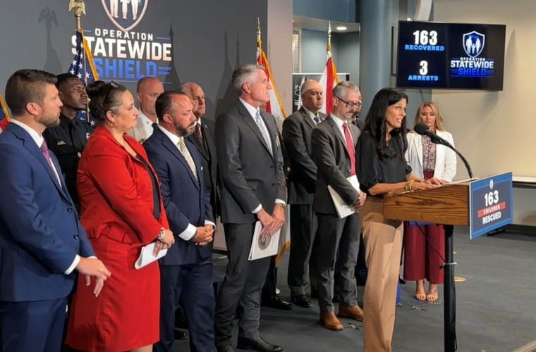 A group of people stands at a podium during a press conference for Operation Statewide Shield. A logo and flags are visible in the background, and a display reads, “163 recovered, 3 arrests.”