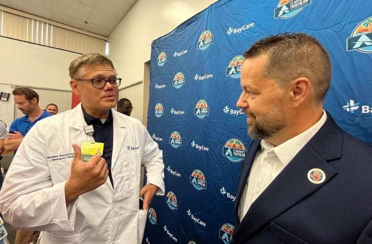 A man in glasses and a doctor's white coat talks with a man wearing sports jacket and a white shirt in front of a blue step and repeat background.