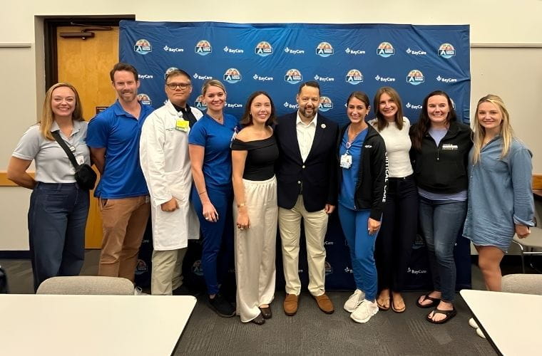 A group of people in street clothes, scrubs and a doctor's jacket pose for a photo inside a large room.