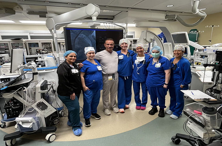 A group of six medical professionals in a hospital setting stands together smiling for a photo. They are surrounded by medical equipment and wearing scrubs and surgical caps. The room is well-lit and appears to be a modern operating or procedure room.