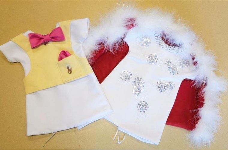 Two handmade infant remembrance outfits are displayed on a table, including a white-and-yellow suit with a pink bow tie and a white gown with silver embellishments and a red velvet cape trimmed with white feathers.