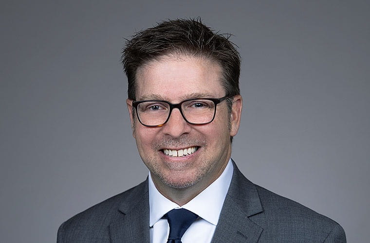 Man with spiky brown hair and brown glasses and wearing a dark gray suit, white shirt and navy-blue tie smiles into the camera.
