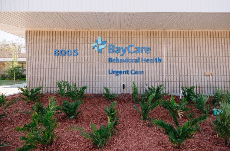 The image shows the exterior of a beige brick building with "BayCare Behavioral Health Urgent Care" and the number "8005" displayed prominently in blue on the wall. In front of the building, there is landscaped red mulch with several green leafy plants.