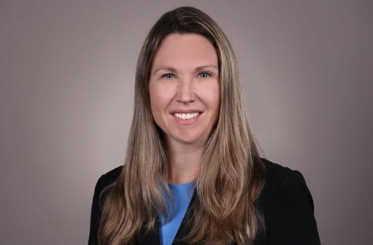 Headshot of Deirdre Franzese, a woman with long light-brown hair wearing a black blazer over a blue top, smiling against a neutral gray background.