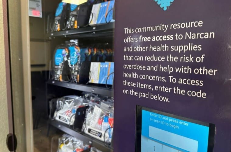 Close-up of a harm reduction vending machine stocked with Narcan and health supplies, with a screen and signage explaining free, anonymous access to overdose prevention resources.