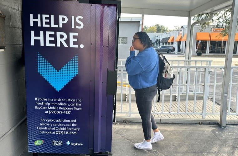 Person stands beside a harm reduction vending machine labeled “Help Is Here,” which provides access to Narcan and addiction recovery resources at a community location.