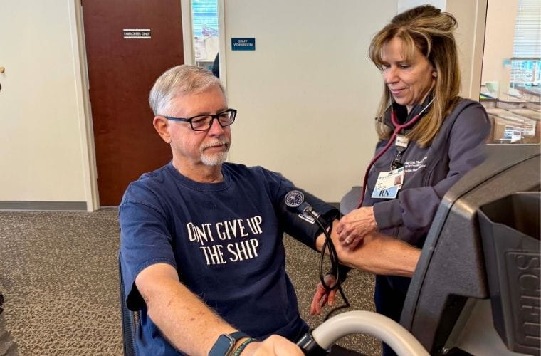 Medical professional in scrubs monitors the blood pressure of a man with gray hair on a recumbent bike.