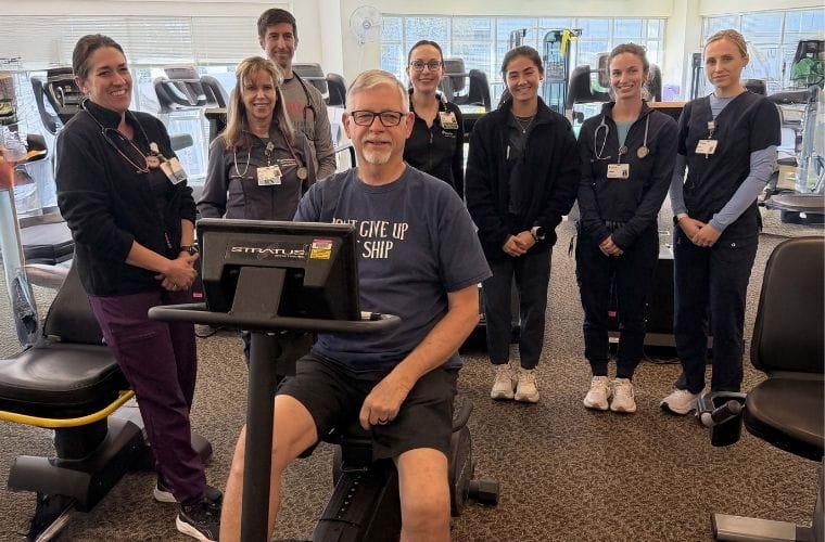 Seven medical professionals in scrubs stand behind a man with gray hair on a recumbent bike.