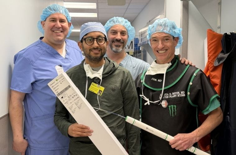 Four men in surgical scrubs standing smiling at the camera. Two in front are holding a box and a long tubular piece of equipment.