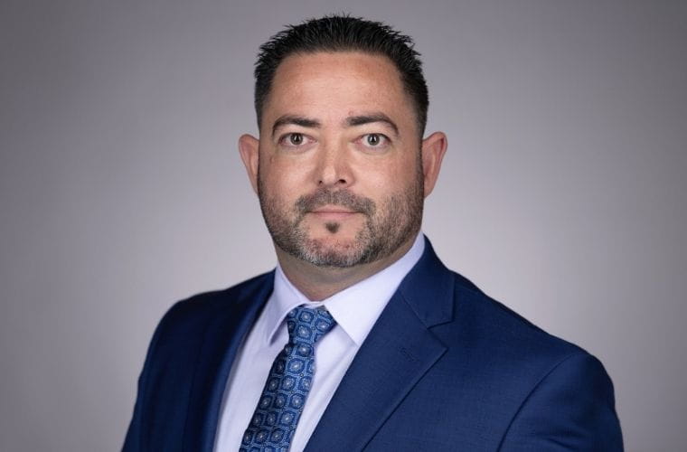 Headshot of Robert Carjaval. He is wearing a navy blue suit, white dress shirt and patterned blue tie, posing for a professional portrait against a plain gray background.