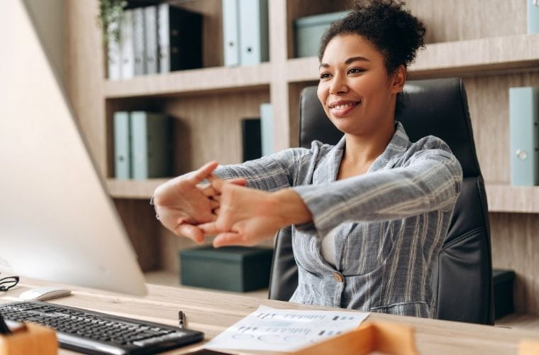 Woman sitting at an office desk stretches her arms forward while looking at a computer screen, with shelves of binders in the background.