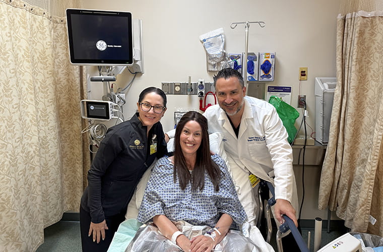 Hospital room scene with a smiling patient in a hospital gown on a bed, surrounded by two doctors. Medical equipment and GE Healthcare monitor visible in the background.