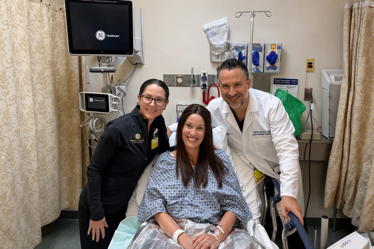 Hospital room scene with a smiling patient in a hospital gown on a bed, surrounded by two doctors. Medical equipment and GE Healthcare monitor visible in the background.