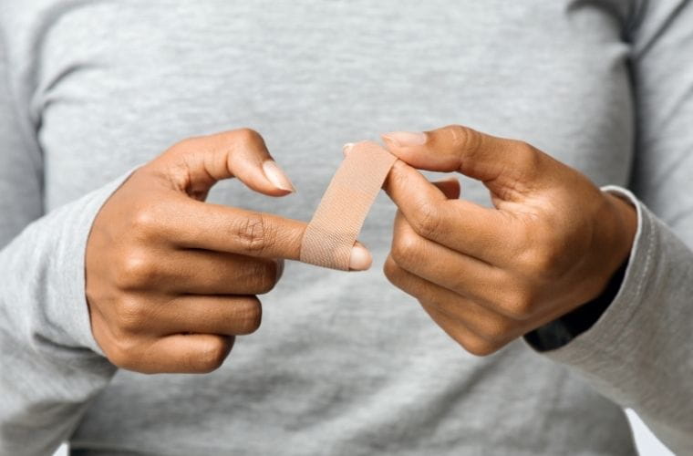  Close-up of a person applying a small adhesive bandage to the tip of a finger, with both hands visible against a neutral background.