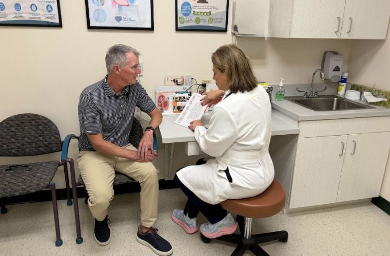 A medical professional in a white coat explains information to a man who is seated in a medical office.