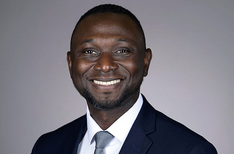 Smiling man with short hair and a short beard wearing a white dress shirt, gray tie and dark suit jacket.