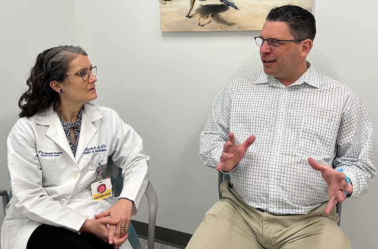 A woman wearing a white physician’s coat talks with a man dressed in a shirt with black lines that form squares and khaki pants in a room with a turtle photo on the wall.
