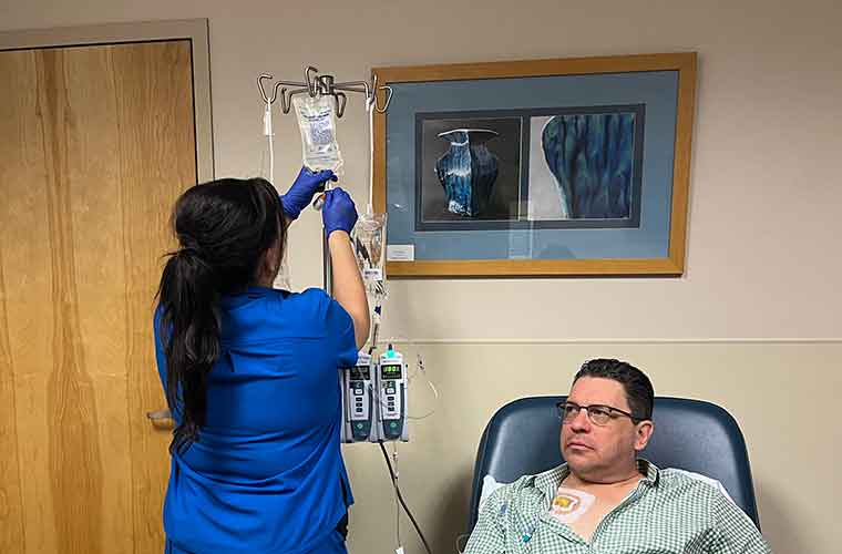 A woman dressed in blue hospital scrubs and blue gloves checks a machine as a man getting a medical procedure sits in a chair. 