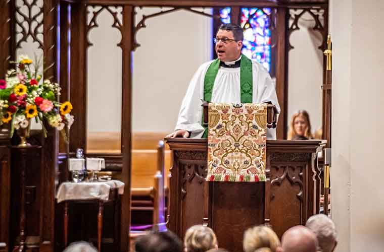 A man dressed in a white religious robe stands in a pulpit and talks to members of a congregation.