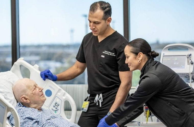 A BayCare Medical Group physician in black scrubs and a clinical nurse in blue care for an elderly patient in around a hospital bed in a hospital room with a view of St. Petersburg.
