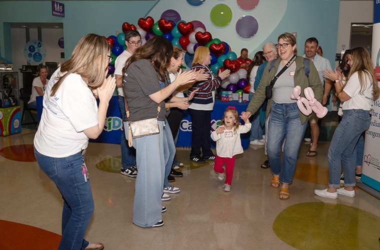 A cheerful gathering in a brightly decorated room with balloons. A child joyfully runs down a path formed by clapping onlookers, holding the hand of an adult, while another holds playful balloon shapes.