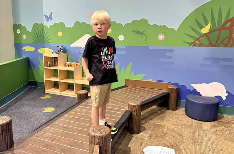 A child with light hair stands on a small wooden bridge in a play area with a nature-themed wall mural. The child wears a black shirt with a graphic saying, "The Little Hearts That Could!" Nearby, there are toys and seating. The setting is colorful and playful.
