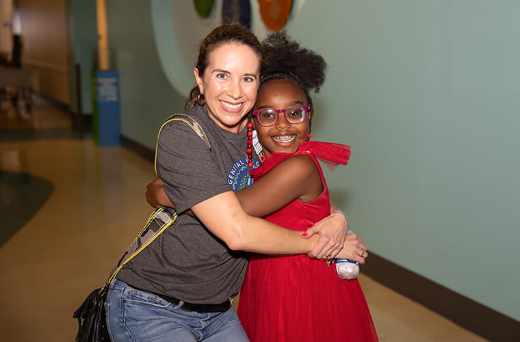 A woman and a child are hugging indoors. One person is wearing a t-shirt and jeans, while the other is in a red dress with matching accessories. The setting appears to be a public space with colorful wall art.