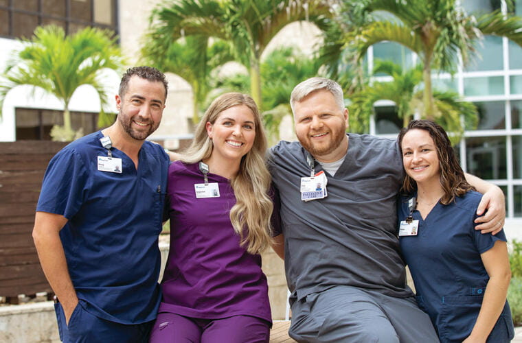 Four BayCare team members in different color scrubs sit together outdoors at a BayCare location. 