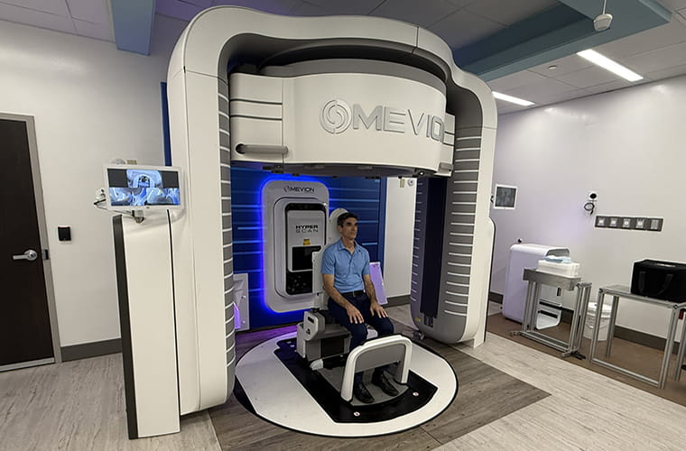 A man wearing a blue polo shirt and black pants sits in a chair inside a piece of medical equipment in a well-lit room.