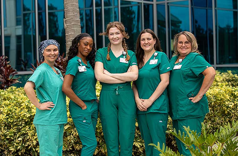 Five smiling women wearing green medical scrubs stand in front of a glass-walled building.